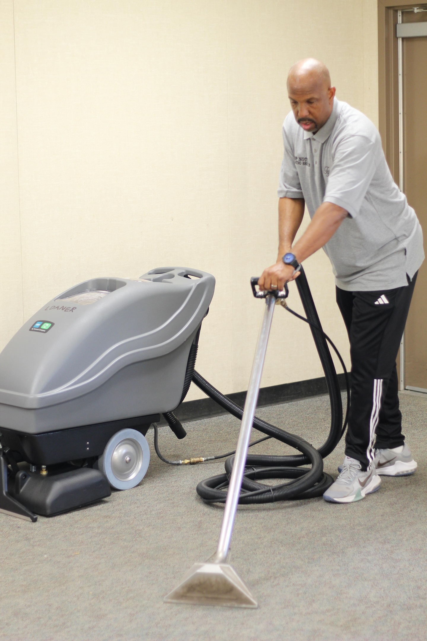 Aerial view of four people in uniform cleaning a modern office space. Two individuals are using mops and a vacuum, while another person is cleaning stairs and the fourth is managing cleaning equipment. The area features plants and contemporary furniture.